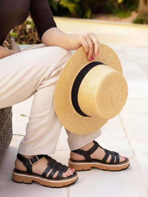 Femme assise portant des sandales noires à brides multiples avec bride cheville ajustable et semelle épaisse, associées à un pantalon beige et un chapeau de paille
