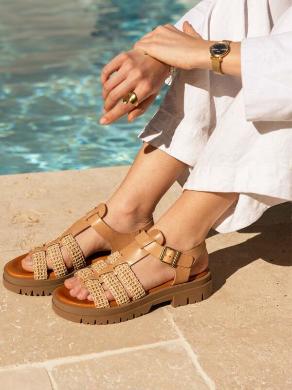 Sandales plates femme en cuir camel avec détails raphia tressé et boucle dorée, collection été 2026, portées au bord d'une piscine avec tenue blanche estivale