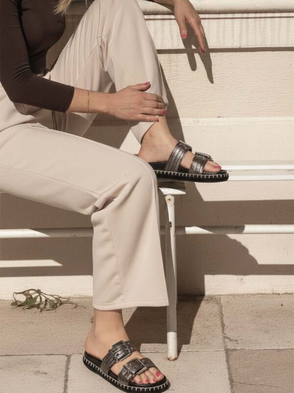 Femme assise portant des mules Semerdjian argentées à double bride avec boucles métalliques et semelle noire contrastée, associées à un pantalon beige