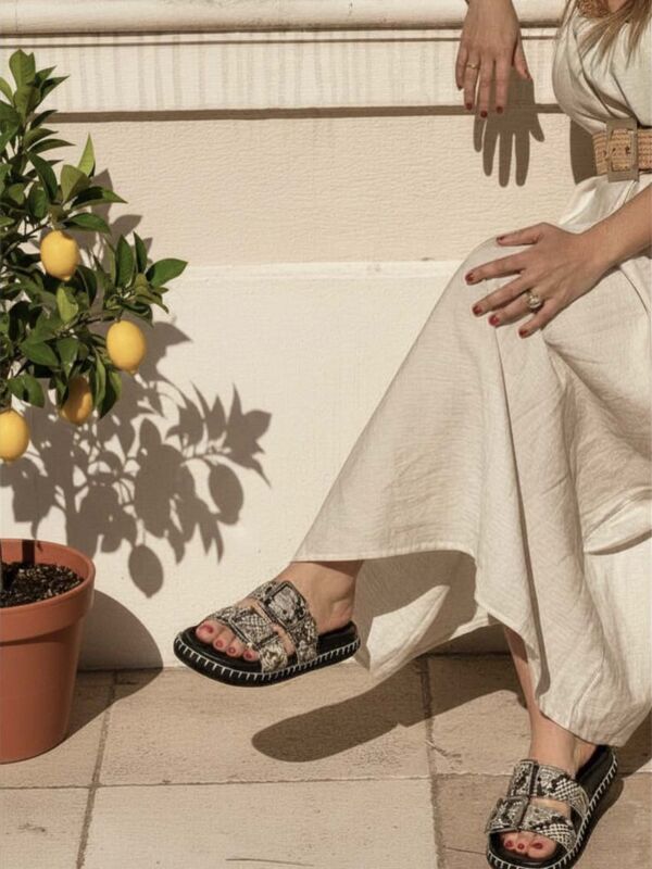 Femme assise portant des mules Semerdjian ou SMR23 ouvertes effet python avec double bride, grande boucle et semelle noire contrastée, associées à une robe longue écrue