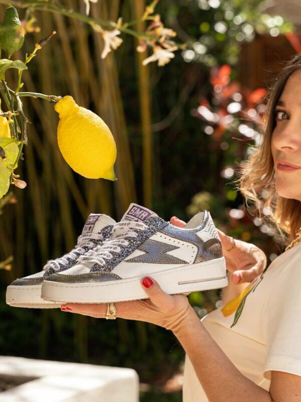 Sneakers SMR23 NOUBAR gris métallisé et paillettes, tenues devant un citronnier. Baskets blanches avec logo argenté brillant et lacets imprimés SMR23.
