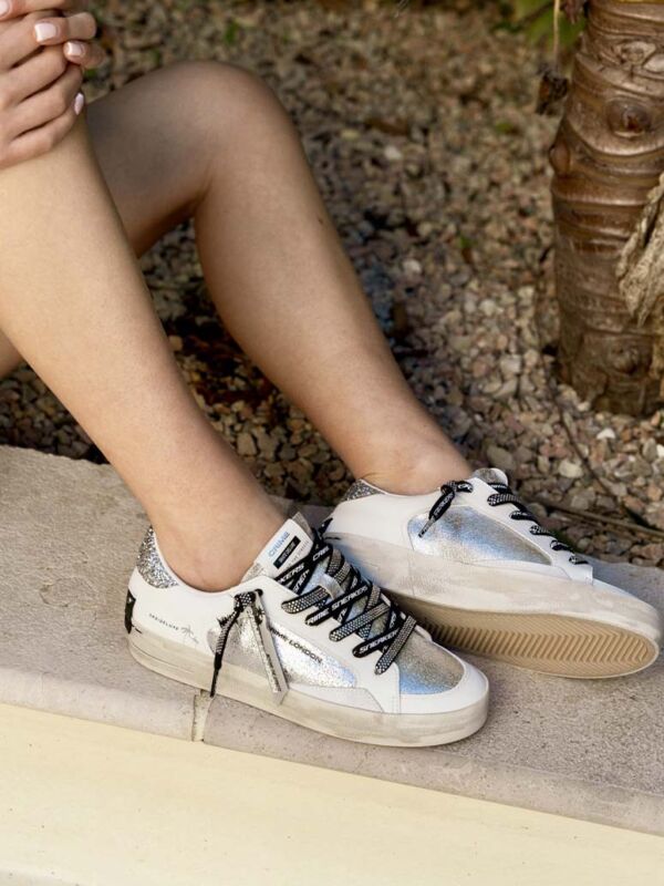 Sneakers femme Crime London blanches avec empiècements argentés scintillants, lacets noirs et blancs siglés et semelle effet vintage, portées en extérieur