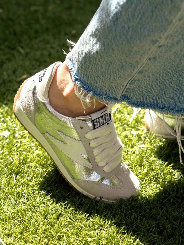 Sneakers Semerdjian June D484 en daim gris métallisé avec logo éclair argenté, lacets blancs texturés et semelle gomme. Gros plan sur la texture cuir brillant et jean effiloché sur pelouse.