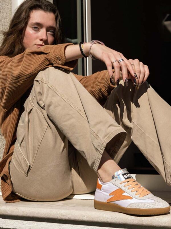 Sneakers femme SMR23 Aslan blanches avec éclair orange, empiècements gris clair, lacets beige et semelle gomme, portées en extérieur