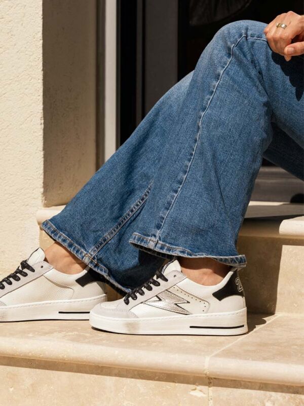 Sneakers blanche Semerdjian en cuir avec lacets noirs et logo éclair argenté métallisé, portée avec un jean taille haute sur des marches ensoleillées.