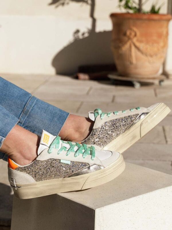 Sneakers femme Crime London compensées blanches avec empiècements pailletés argentés, lacets verts d’eau et détail orange au talon, portées en extérieur