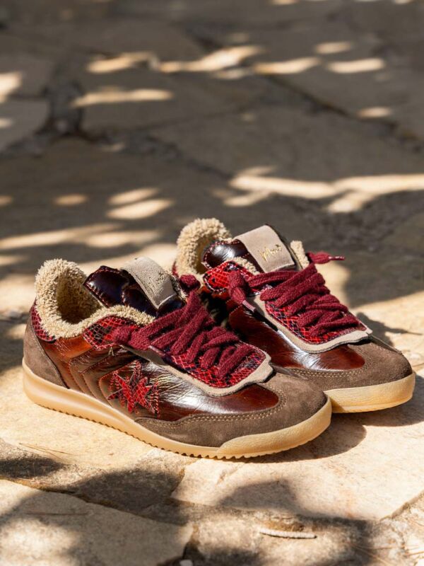 Paire de sneakers Moni Shoes LOVLAF502 marron bordeaux posée sur pierre en extérieur, lumière naturelle avec ombres de feuilles, col teddy apparent et semelle gomme.